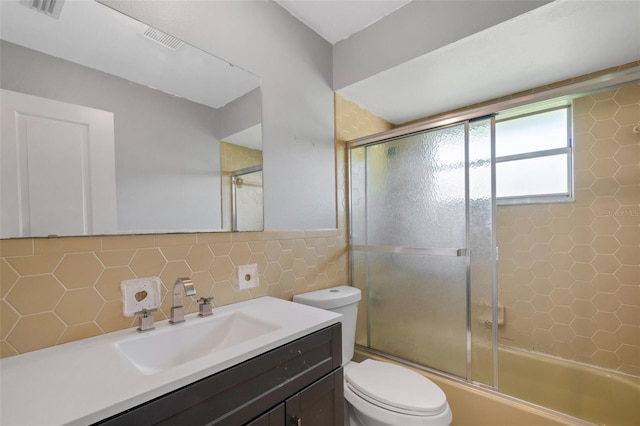 bathroom with toilet, visible vents, vanity, tile walls, and combined bath / shower with glass door