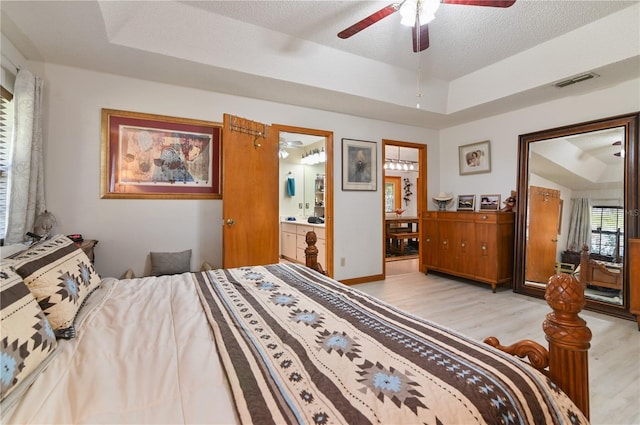 bedroom with connected bathroom, ceiling fan, light hardwood / wood-style flooring, a textured ceiling, and a tray ceiling