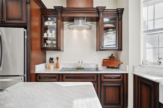 kitchen featuring black electric cooktop, wall chimney range hood, freestanding refrigerator, light stone countertops, and glass insert cabinets