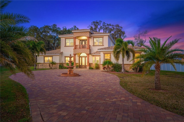 mediterranean / spanish home featuring a balcony, stone siding, decorative driveway, french doors, and stucco siding