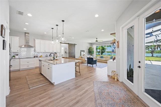 kitchen with an island with sink, white cabinets, wall chimney exhaust hood, light hardwood / wood-style flooring, and ceiling fan