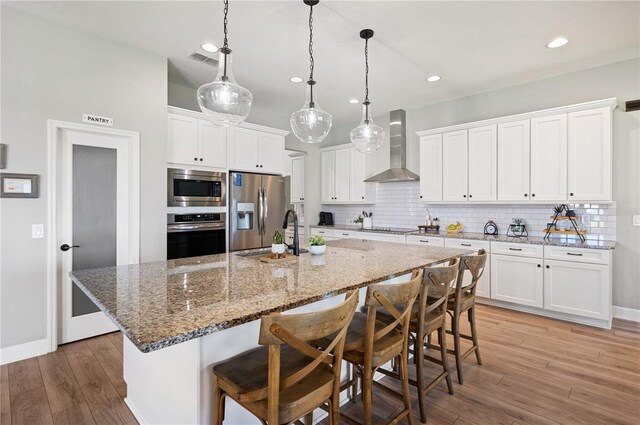 kitchen featuring white cabinets, a large island, wall chimney range hood, stainless steel appliances, and light hardwood / wood-style floors