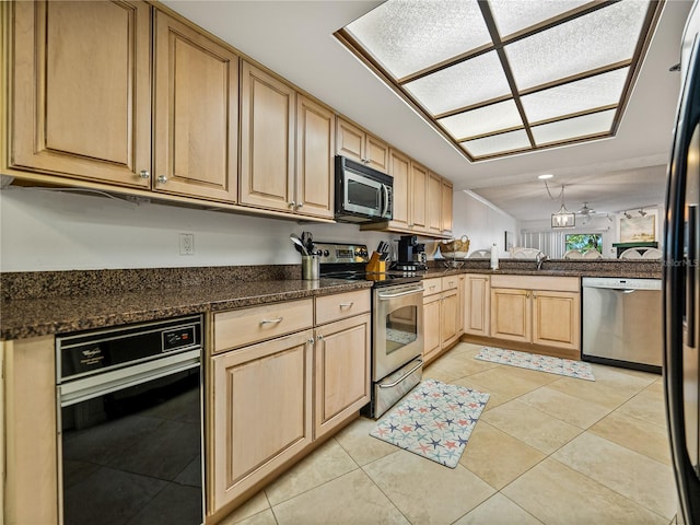 kitchen with light brown cabinetry, kitchen peninsula, appliances with stainless steel finishes, light tile floors, and dark stone countertops