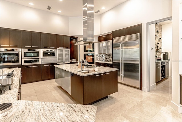 kitchen featuring island range hood, a high ceiling, appliances with stainless steel finishes, dark brown cabinets, and an island with sink