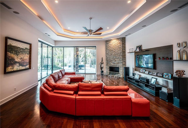 living room with dark hardwood / wood-style flooring, ceiling fan, a stone fireplace, and a tray ceiling