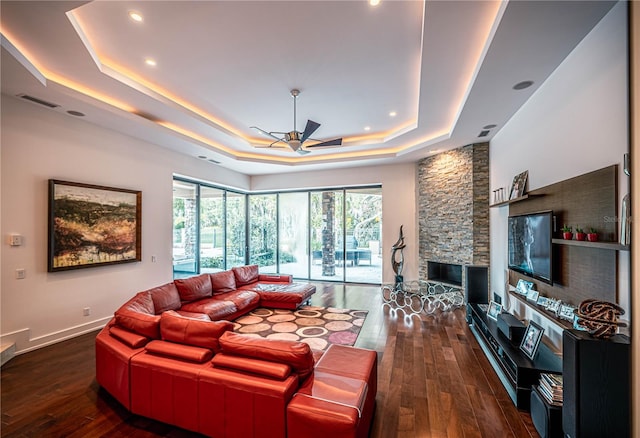 living room featuring ceiling fan, dark wood-type flooring, a tray ceiling, and a stone fireplace