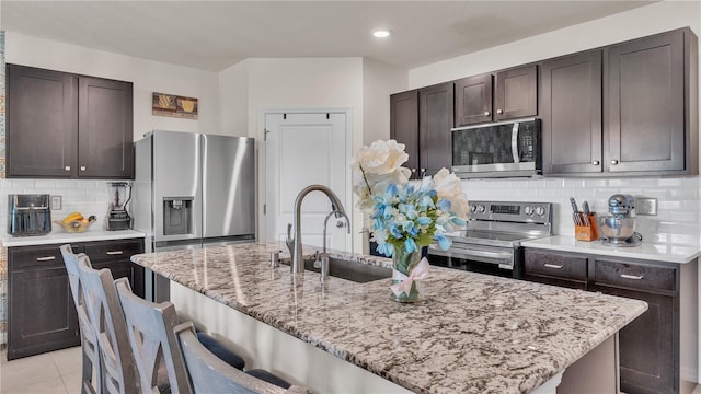 kitchen with stainless steel appliances, an island with sink, backsplash, sink, and light tile floors