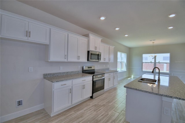 kitchen with white cabinetry, stainless steel appliances, a kitchen island with sink, a sink, and light stone countertops
