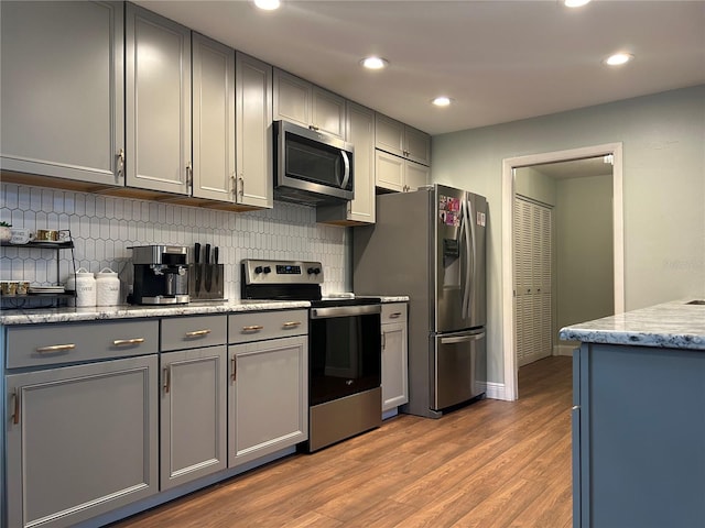 kitchen with decorative backsplash, appliances with stainless steel finishes, light stone counters, gray cabinetry, and hardwood / wood-style flooring