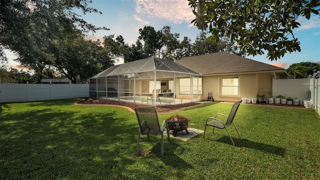 back house at dusk featuring glass enclosure, a fire pit, and a lawn