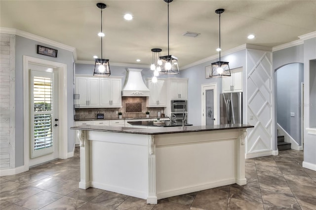 kitchen with a center island with sink, hanging light fixtures, custom range hood, white cabinetry, and stainless steel appliances