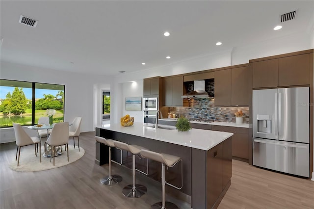 kitchen featuring a center island with sink, stainless steel appliances, tasteful backsplash, light wood-type flooring, and wall chimney exhaust hood