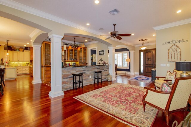 living room with ornate columns, ceiling fan with notable chandelier, crown molding, indoor bar, and hardwood / wood-style flooring