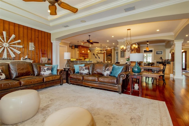 living room with ornate columns, dark wood-type flooring, a raised ceiling, ceiling fan with notable chandelier, and ornamental molding