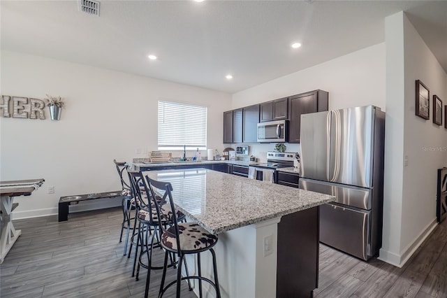 kitchen with appliances with stainless steel finishes, hardwood / wood-style flooring, light stone countertops, a center island, and a breakfast bar