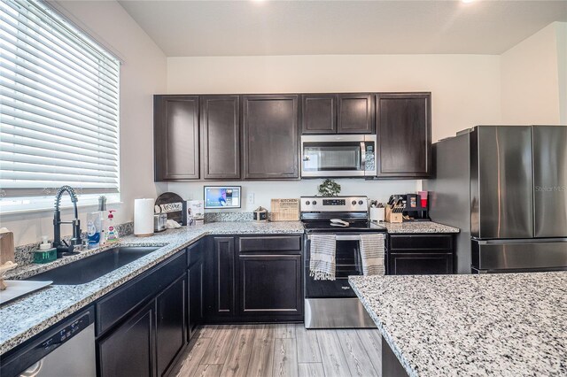 kitchen with appliances with stainless steel finishes, dark brown cabinets, light stone countertops, sink, and light hardwood / wood-style flooring