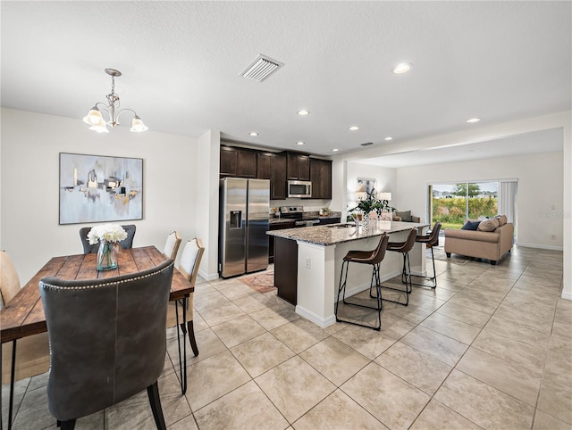 kitchen with stainless steel appliances, an island with sink, light stone countertops, dark brown cabinetry, and decorative light fixtures