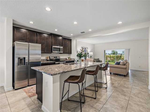 kitchen featuring light stone counters, dark brown cabinets, a center island with sink, a breakfast bar, and appliances with stainless steel finishes