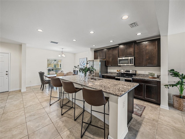 kitchen featuring light stone counters, stainless steel appliances, a kitchen bar, a kitchen island with sink, and dark brown cabinets