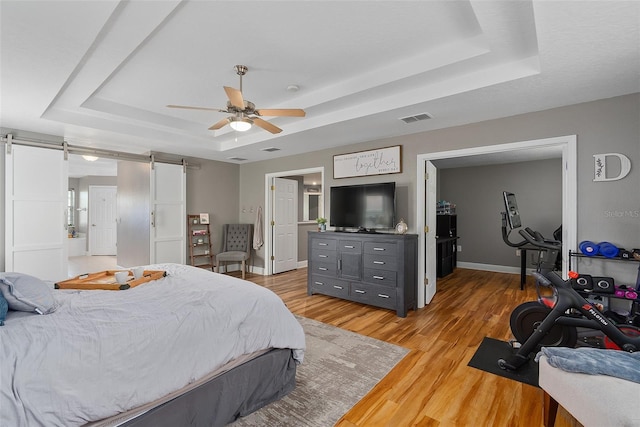 bedroom with light wood-type flooring, ceiling fan, a barn door, and a tray ceiling