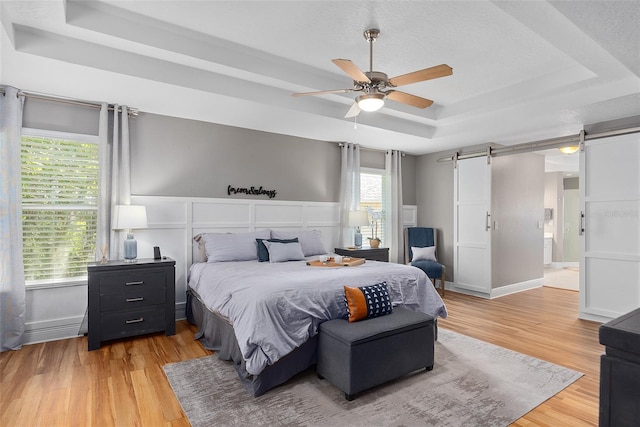 bedroom with ensuite bath, a raised ceiling, a barn door, light wood-type flooring, and ceiling fan