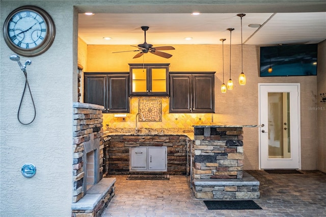 kitchen with sink, light stone countertops, ceiling fan, a stone fireplace, and dark brown cabinetry