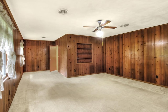 empty room featuring wood walls, ornamental molding, built in features, ceiling fan, and light colored carpet