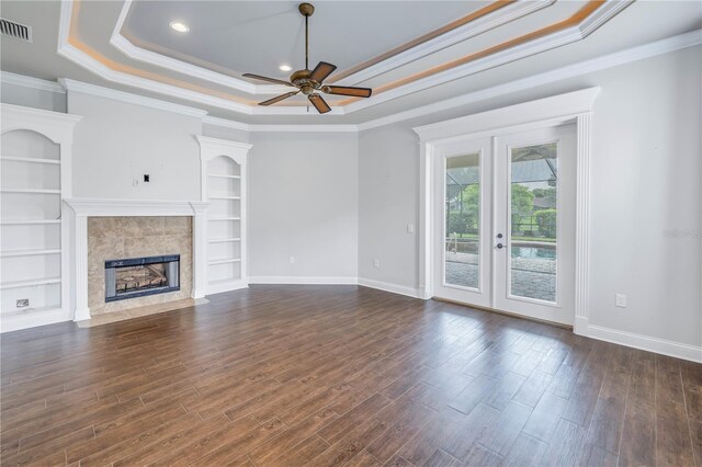 unfurnished living room featuring ceiling fan, built in shelves, a raised ceiling, a tiled fireplace, and dark wood-type flooring