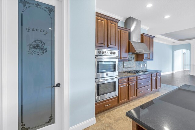 kitchen with wall chimney exhaust hood, stainless steel appliances, backsplash, light tile patterned floors, and crown molding