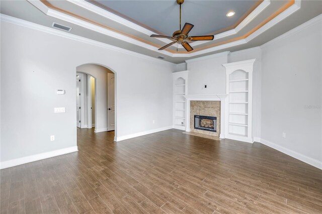 unfurnished living room with wood-type flooring, a tray ceiling, a tile fireplace, built in shelves, and ceiling fan