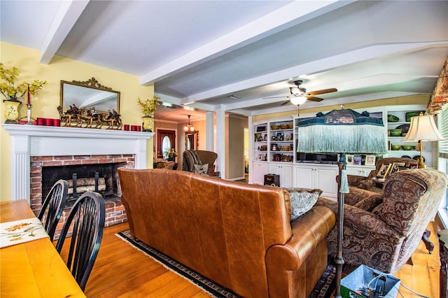 living room featuring beamed ceiling, light wood-type flooring, a brick fireplace, and ceiling fan with notable chandelier