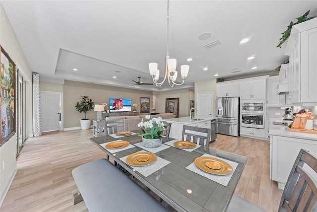 dining area with ceiling fan with notable chandelier, light hardwood / wood-style floors, sink, and a tray ceiling