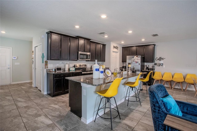 kitchen featuring dark brown cabinetry, an island with sink, appliances with stainless steel finishes, a breakfast bar area, and dark stone counters