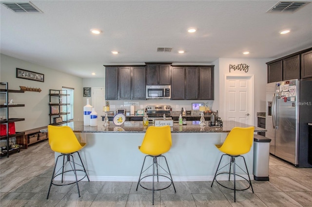 kitchen featuring a breakfast bar, a kitchen island with sink, appliances with stainless steel finishes, dark brown cabinetry, and dark stone countertops