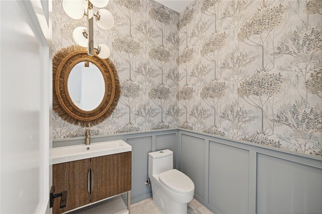bathroom featuring vanity, toilet, wainscoting, a decorative wall, and wallpapered walls