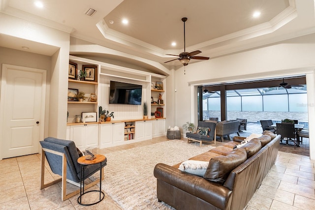 living room featuring built in shelves, light tile patterned floors, ornamental molding, ceiling fan, and a tray ceiling