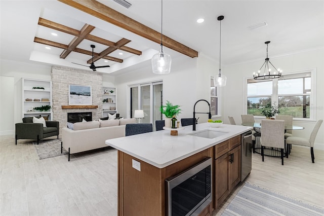 kitchen with beam ceiling, a kitchen island with sink, light wood-type flooring, sink, and decorative light fixtures