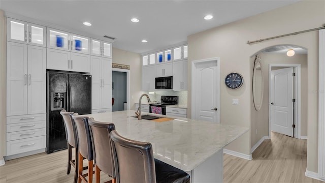 kitchen with light stone countertops, black appliances, a center island with sink, light hardwood / wood-style floors, and white cabinetry