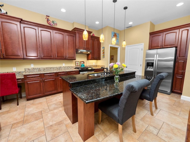 kitchen featuring stainless steel appliances, hanging light fixtures, a kitchen island with sink, a breakfast bar, and dark stone countertops