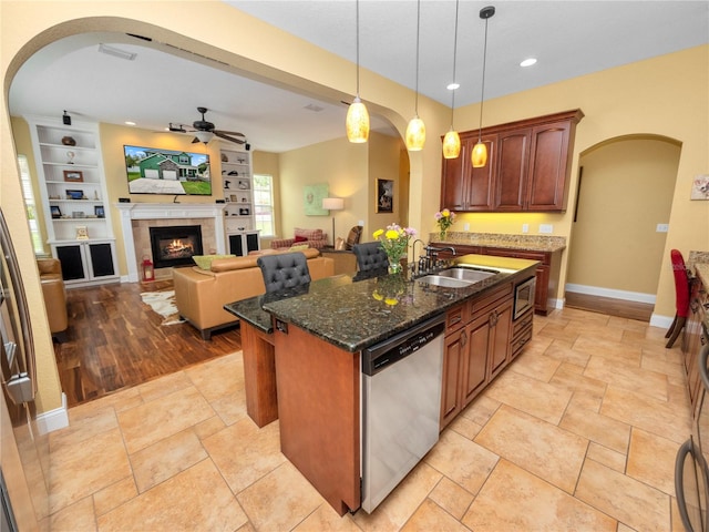 kitchen with sink, dark stone counters, hanging light fixtures, an island with sink, and dishwasher