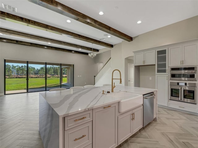 kitchen featuring a kitchen island with sink, sink, beam ceiling, light stone counters, and stainless steel appliances