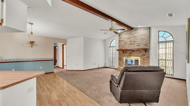 living room with a brick fireplace, a textured ceiling, light colored carpet, ceiling fan, and lofted ceiling with beams