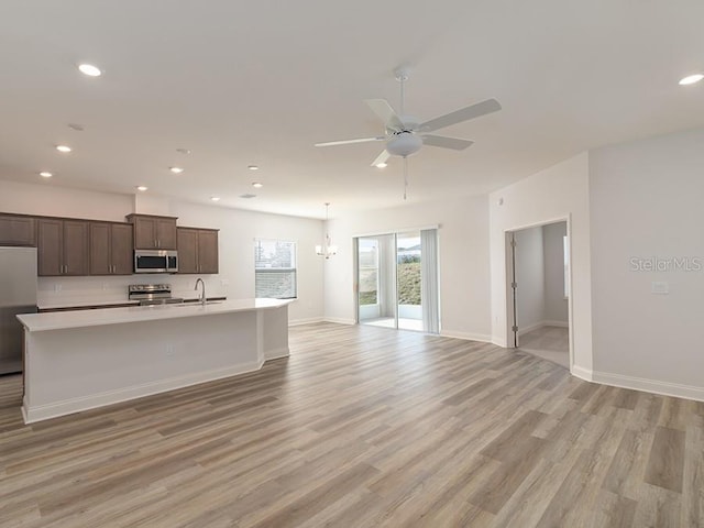 kitchen with appliances with stainless steel finishes, ceiling fan with notable chandelier, dark brown cabinetry, a center island with sink, and light wood-type flooring