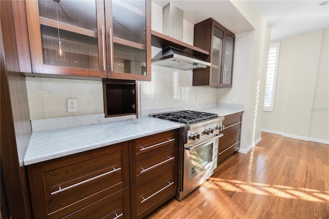 kitchen with decorative backsplash, light wood-type flooring, light stone counters, high end stove, and wall chimney range hood