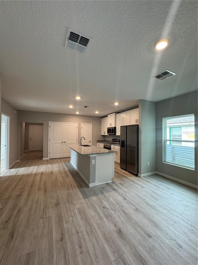 kitchen featuring appliances with stainless steel finishes, light wood-type flooring, light stone counters, white cabinetry, and an island with sink