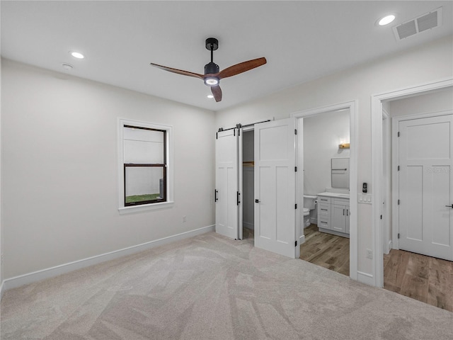 unfurnished bedroom featuring ceiling fan, a barn door, light colored carpet, and connected bathroom