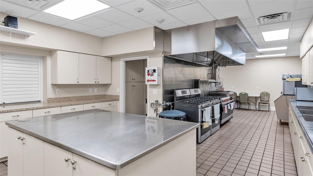 kitchen with a center island, a drop ceiling, wall chimney range hood, stainless steel gas range, and white cabinets