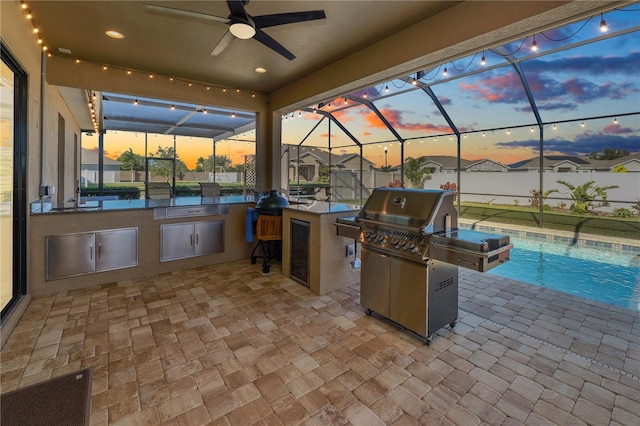 patio terrace at dusk featuring ceiling fan, an outdoor kitchen, a lanai, grilling area, and a fenced in pool