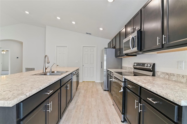 kitchen with vaulted ceiling, appliances with stainless steel finishes, sink, a kitchen island with sink, and light wood-type flooring