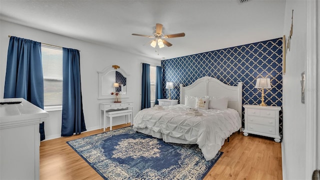 bedroom featuring wallpapered walls, an accent wall, baseboards, ceiling fan, and light wood-style flooring